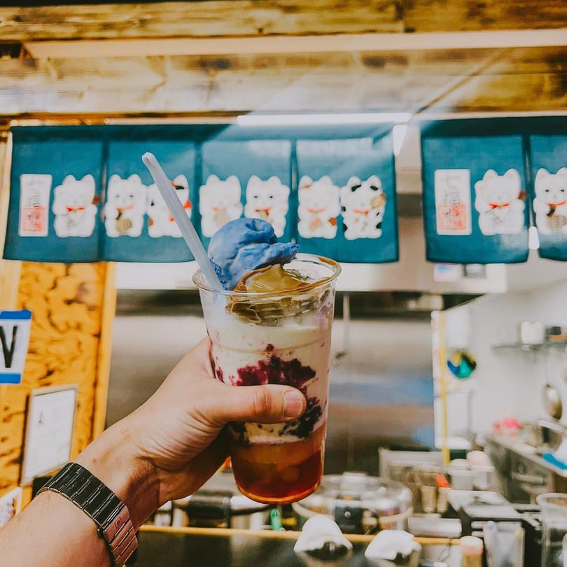 Hand holding a layered dessert drink with blue ice cream, caramel, and berries, in front of Japanese lucky cat banners.